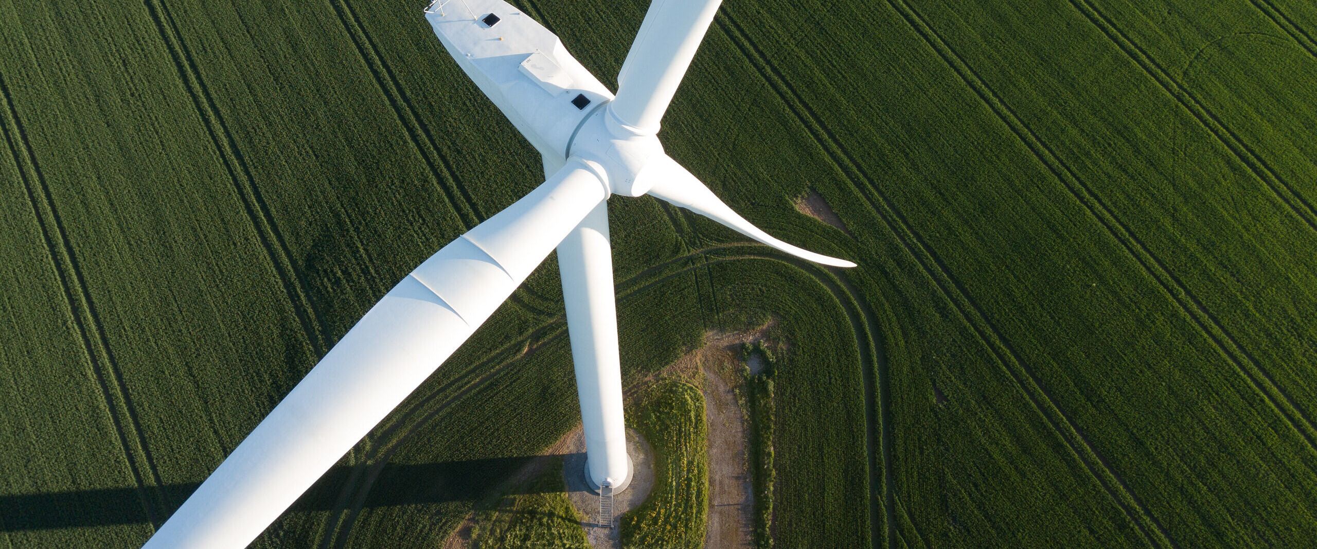 A close-up aerial view of a white wind turbine surrounded by lush green fields. The turbine's blades are captured at an angle, providing a glimpse of its large size. The field is marked with tractor lines, creating a textured landscape.