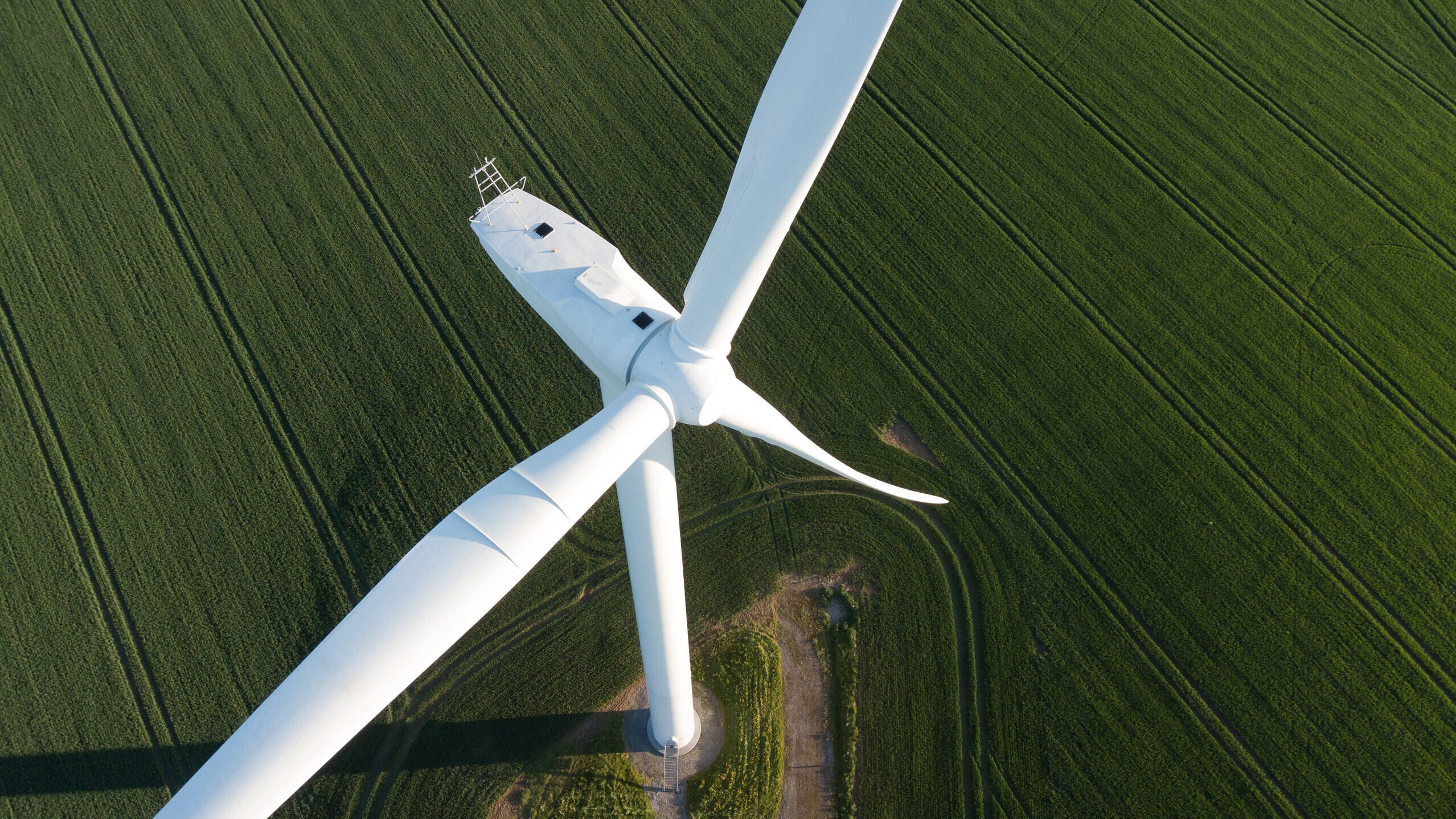 A close-up aerial view of a white wind turbine surrounded by lush green fields. The turbine's blades are captured at an angle, providing a glimpse of its large size. The field is marked with tractor lines, creating a textured landscape.
