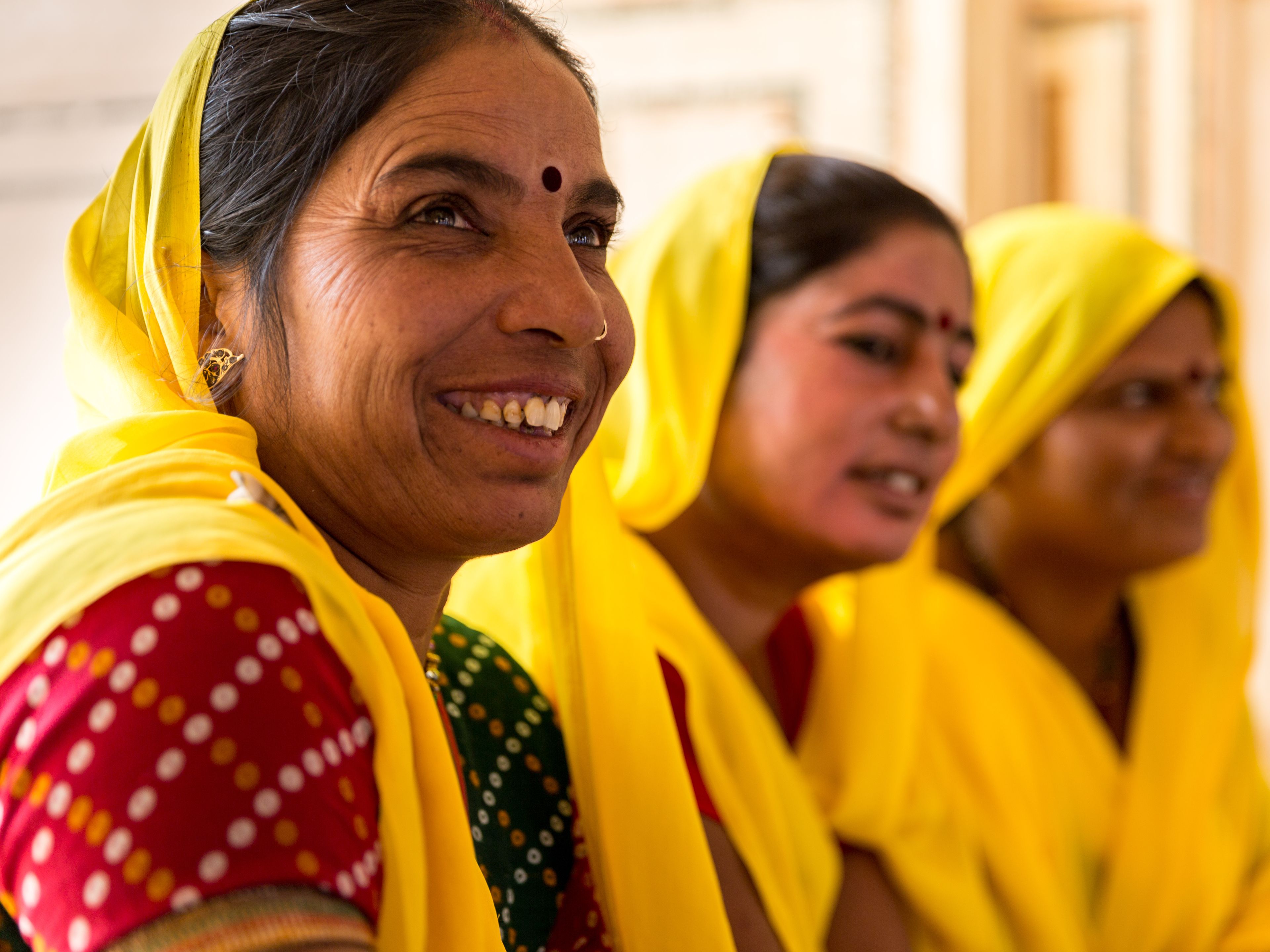 Three women dressed in vibrant yellow sarees and traditional attire are gathered together, smiling and conversing. The woman in the foreground has a joyful expression and a red bindi on her forehead. The background is softly blurred, highlighting their interaction.